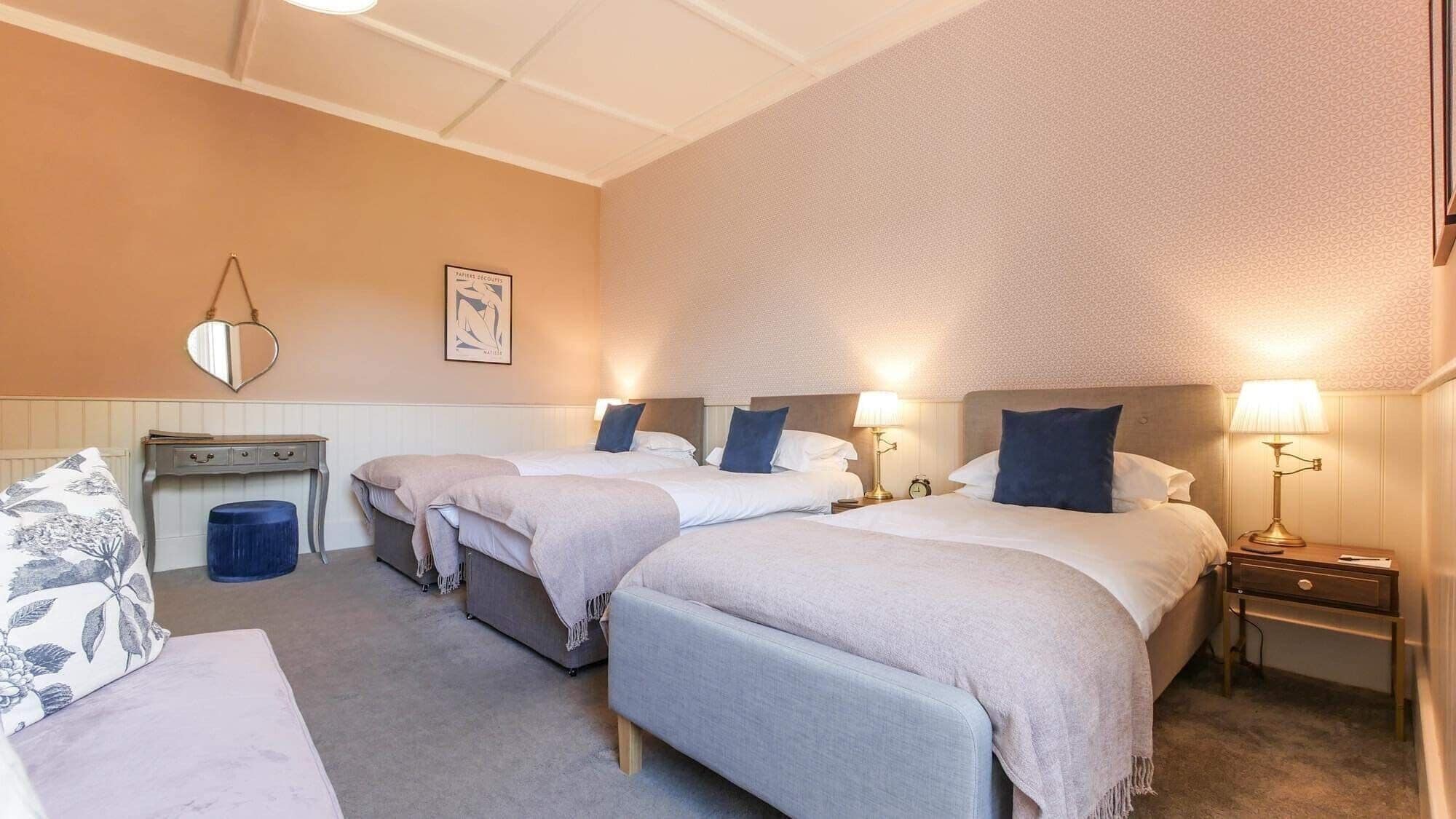 Une chambre d’hôtel avec trois lits simples, un petit bureau, une chaise et une table de chevet avec une lampe.