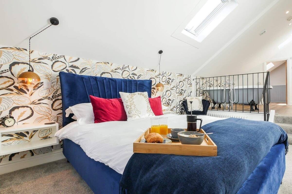 A bedroom with a bed, a tray with breakfast items, a clock, and a window with a view of a bathtub.