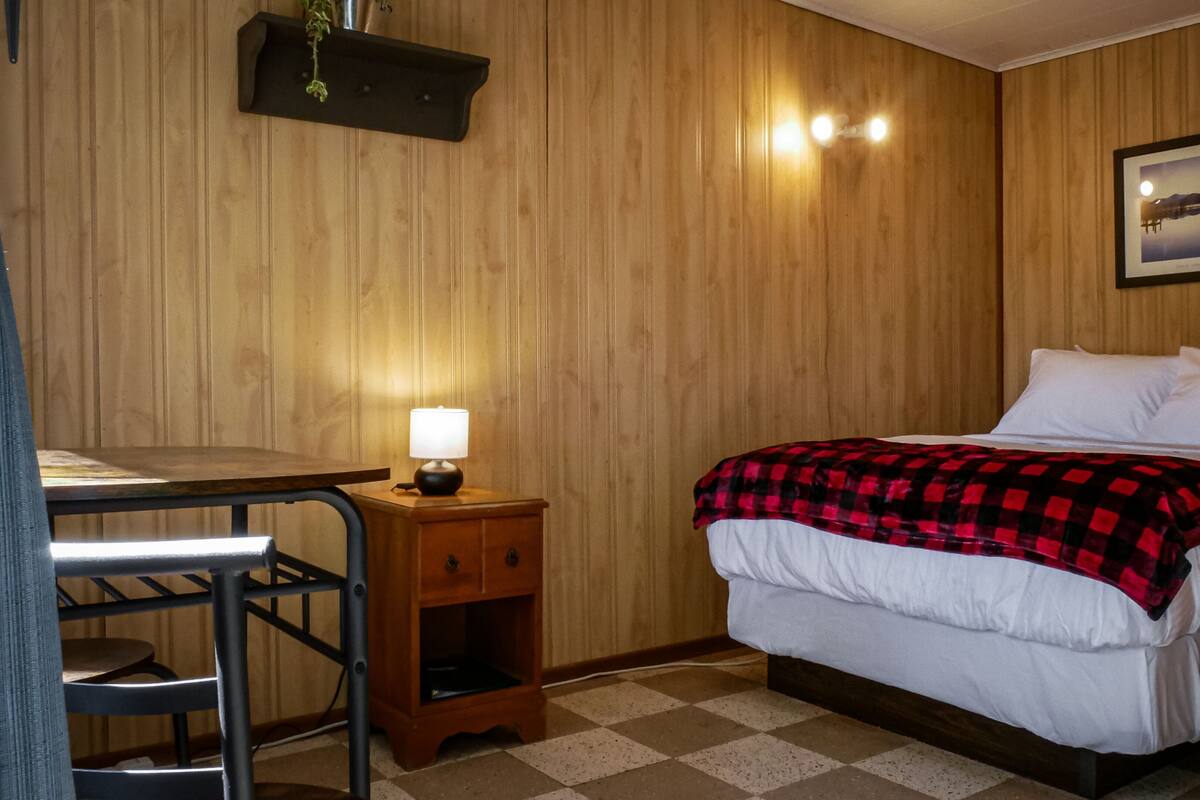 Una habitación con una cama, un escritorio, una silla, una mesita de noche con lámpara y un cuadro en la pared.