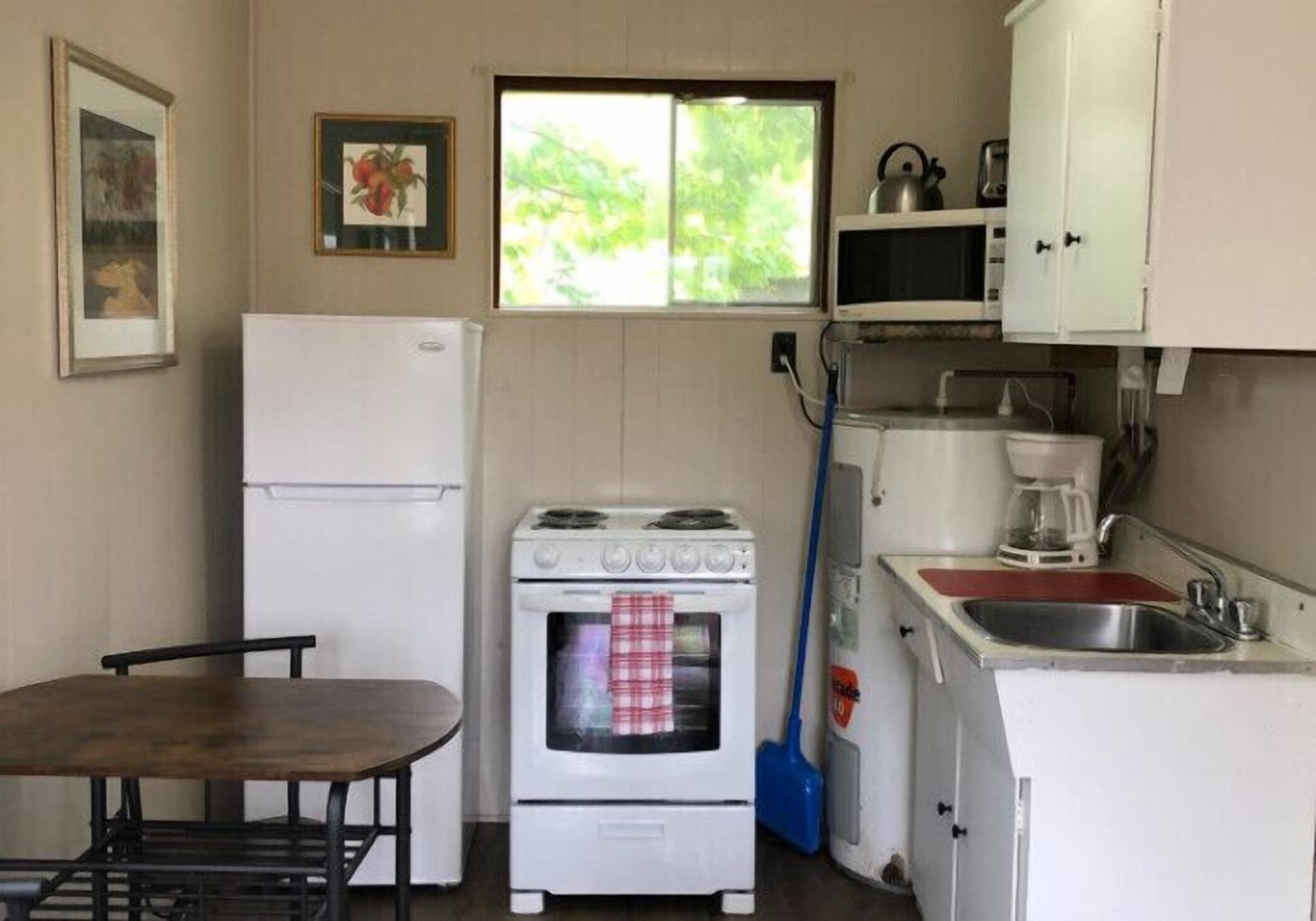 Eine kleine Küche mit Kühlschrank, Herd, Mikrowelle und Spüle. Ein Fenster sorgt für natürliches Licht, und ein Besen lehnt an der Wand.