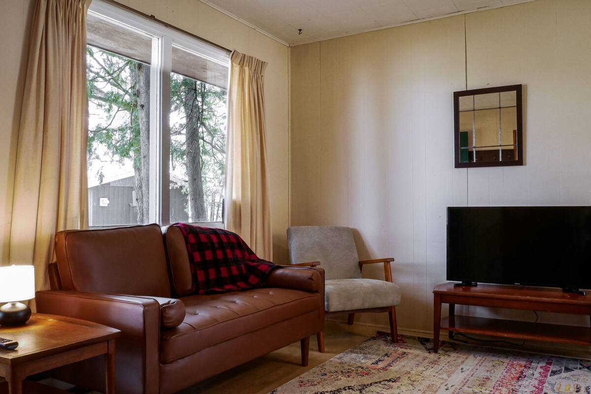 Una sala de estar con un sofá de cuero marrón, un sillón blanco, un televisor sobre un mueble de madera y una ventana con cortinas.