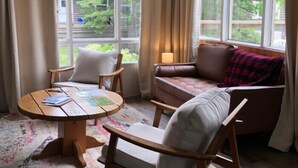 Una sala de estar acogedora con un sofá de cuero marrón, una mesa de centro de madera y dos sillones blancos. Hay una lámpara sobre el sofá y la habitación tiene ventanas grandes con cortinas.