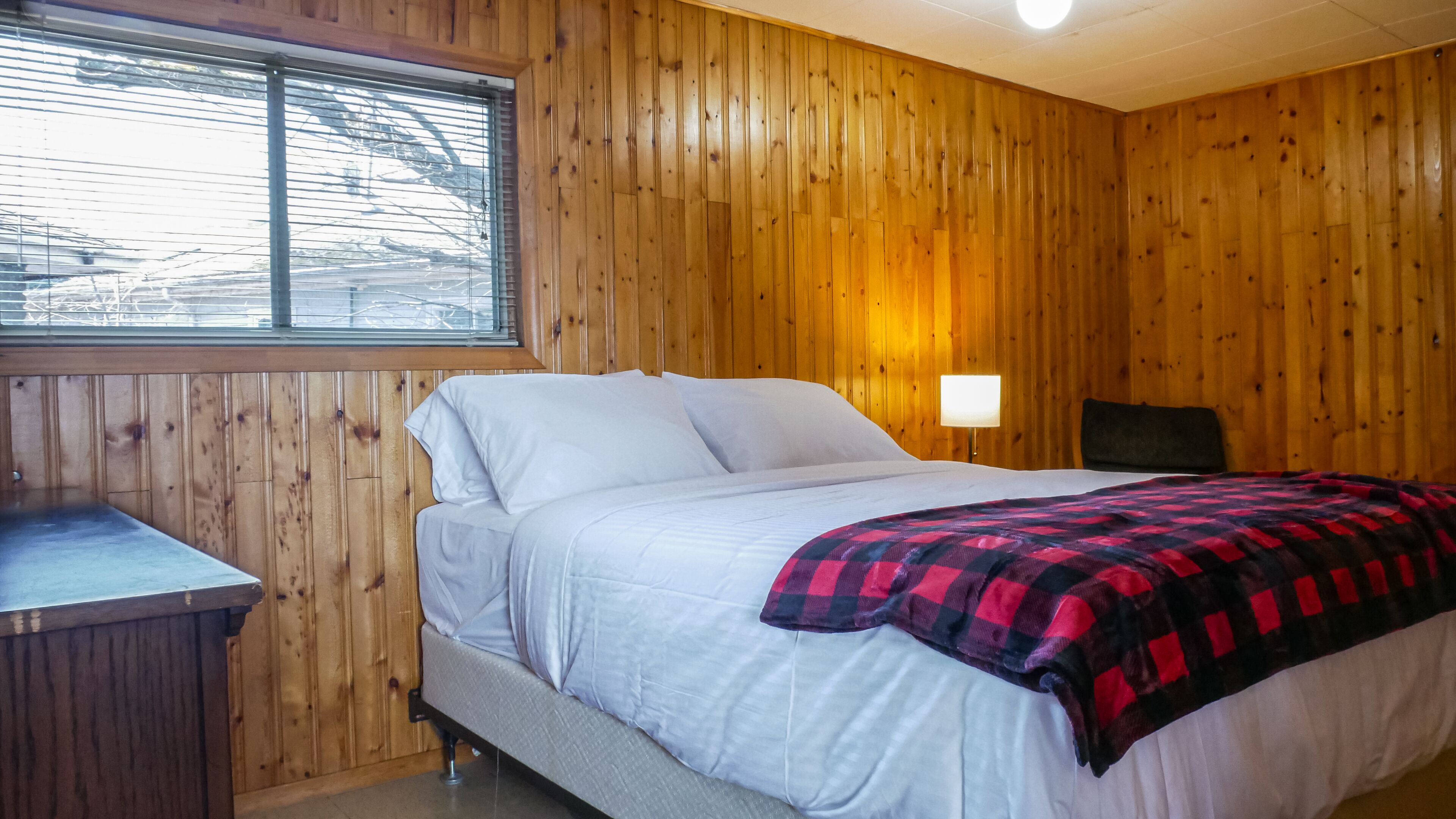 Una habitación con paneles de madera, una cama, una mesita de noche y un ventanal con persianas.