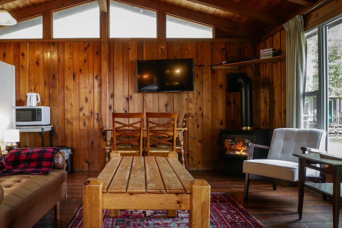 Un acogedor interior de cabaña con paredes de madera, un televisor montado en la pared, una chimenea, un sofá y una mesa de comedor.