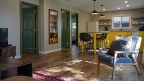 Una sala de estar moderna con un sofá, una mesa de centro y un televisor. Hay una zona de cocina con un refrigerador, un lavamanos y una ventana.