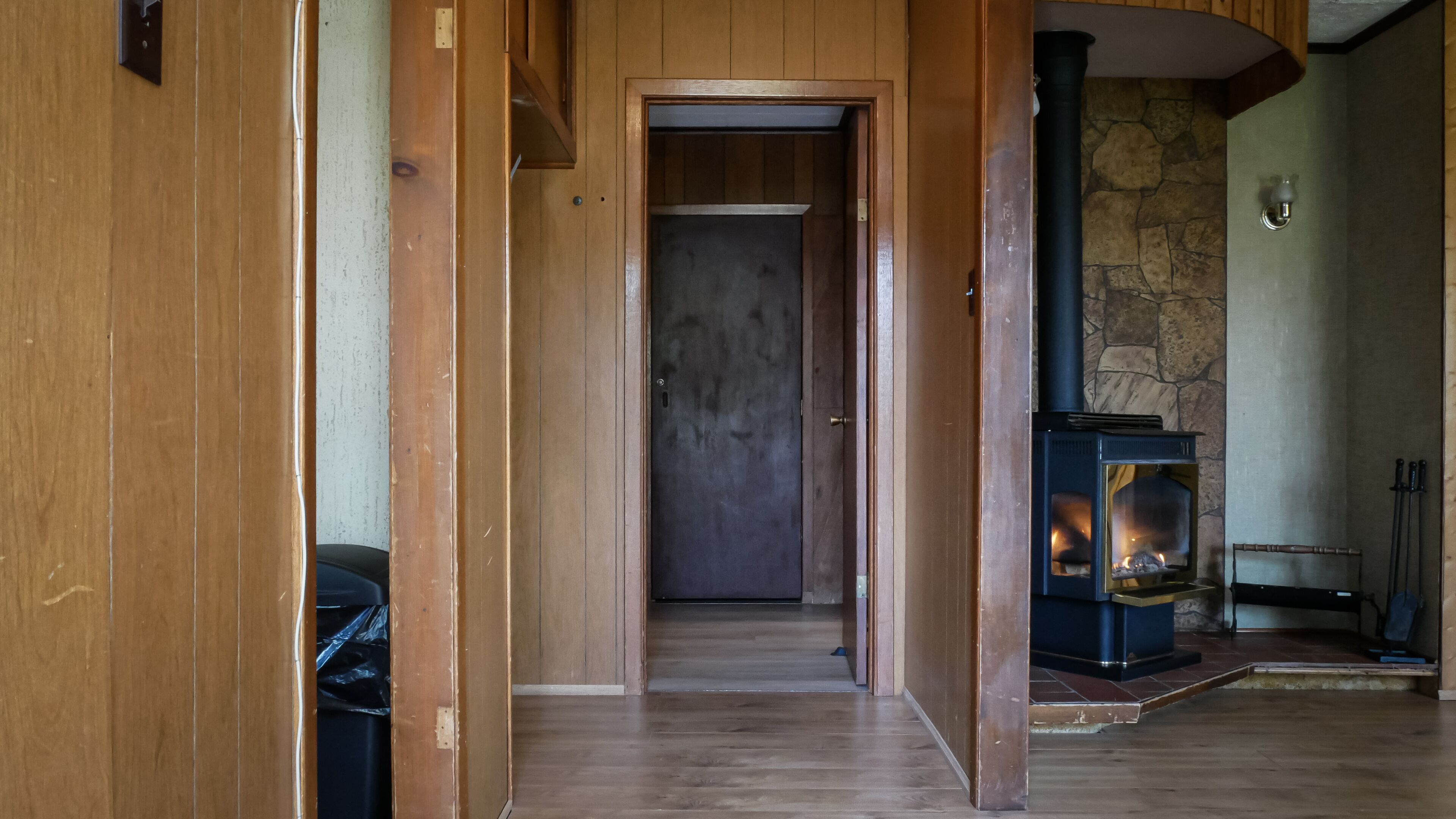 Un couloir en bois mène à une pièce dont un des murs est en pierre et qui est équipée d’un poêle à bois.