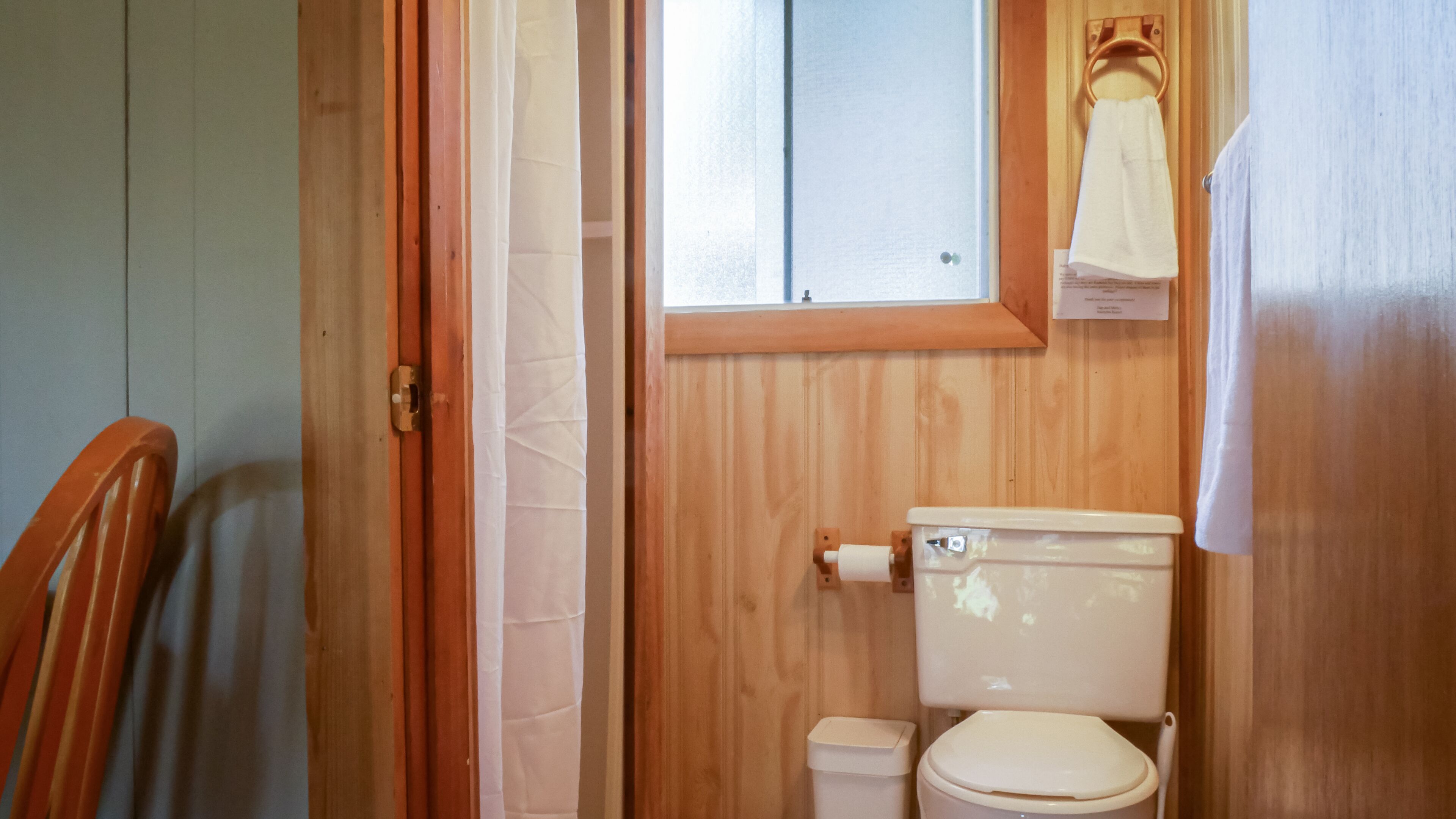 Un baño pequeño con paneles de madera, un inodoro, una ventana y una toalla colgada en un perchero.