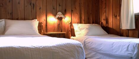 A room with two beds, wooden walls, and a nightstand with a lamp.