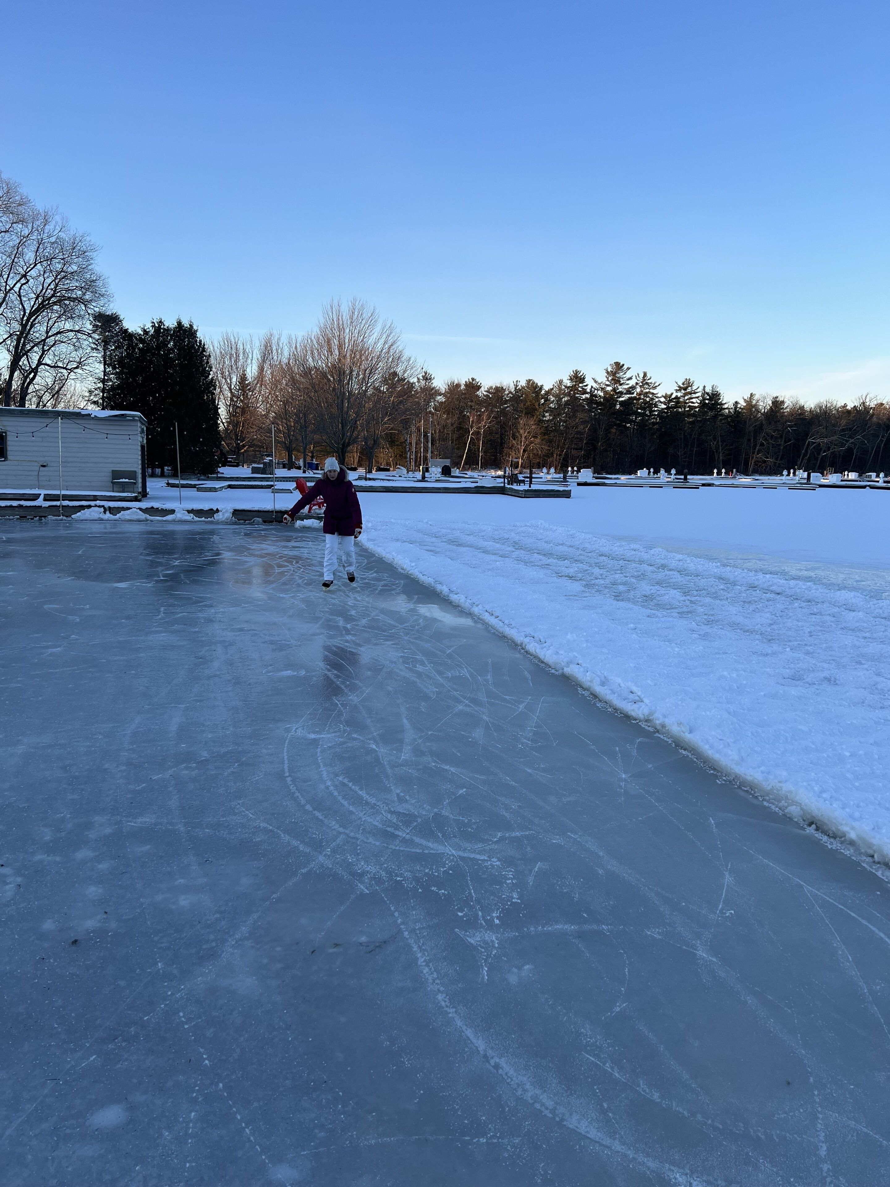 A frozen outdoor area with a person skating, a trailer, and trees in the background.