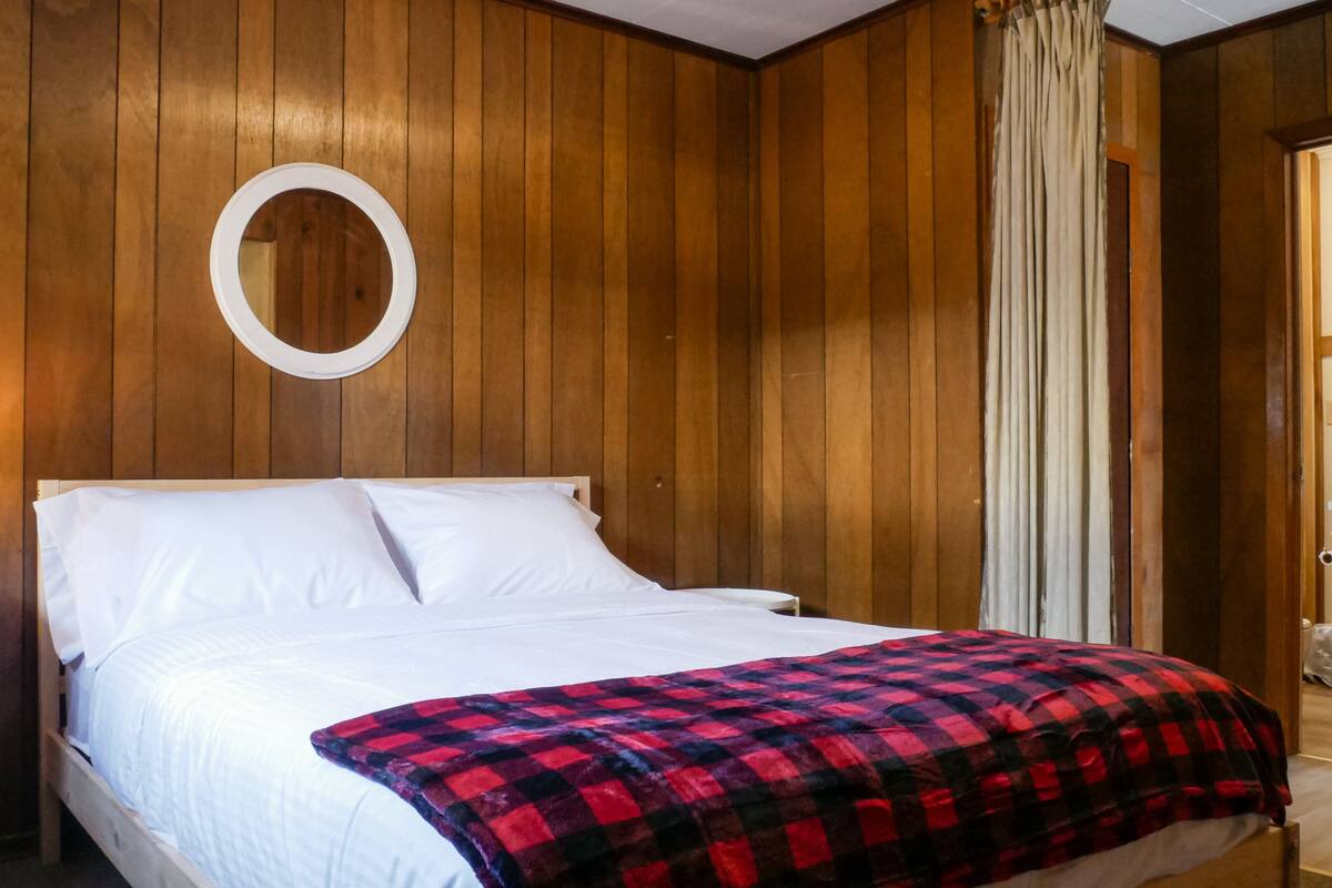 Un dormitorio con un revestimiento de madera, un espejo redondo, una cama con ropa de cama blanca, una manta de cuadros rojos y negros y una mesita de noche con una lámpara.
