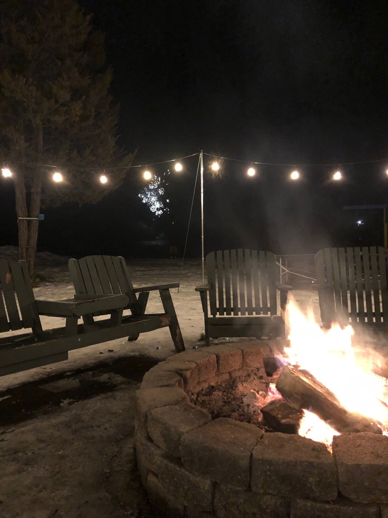 A nighttime outdoor seating area with two Adirondack chairs, a fire pit, and string lights.