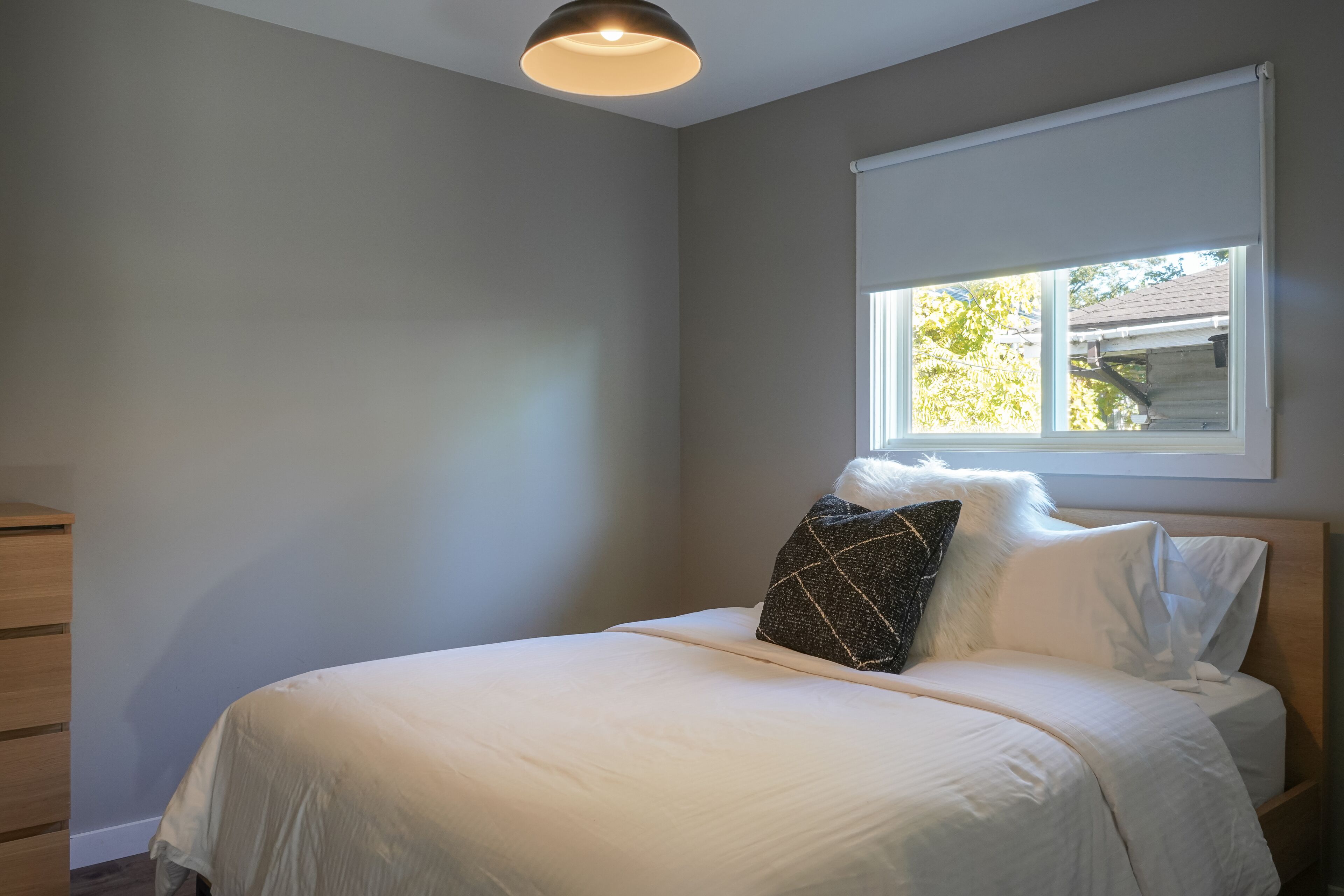 A bedroom with a bed, a window with blinds, a ceiling light, and a wooden dresser.