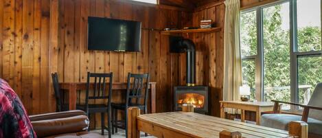 Interior acogedor de una cabaña con paredes de madera, una chimenea, un televisor montado en la pared y un ventanal con vistas al exterior.