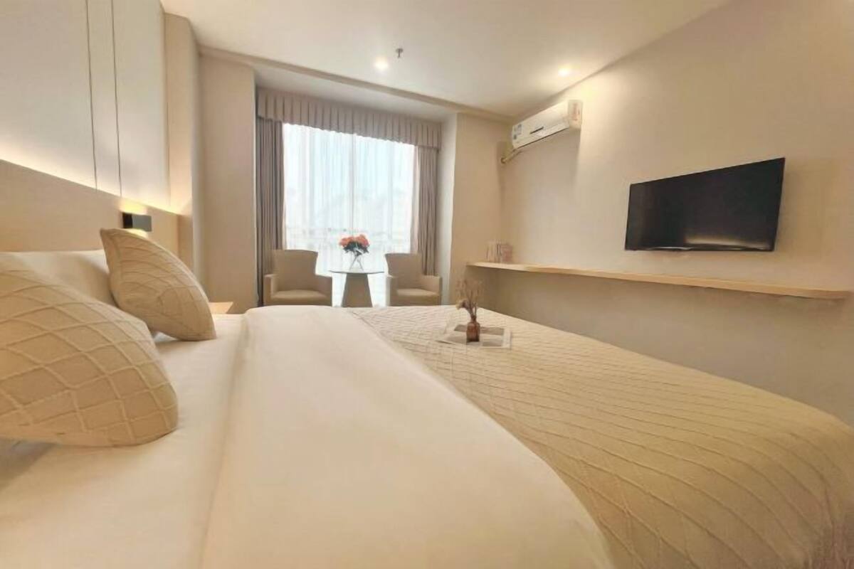 A hotel room with a bed, a TV mounted on the wall, a small table with a vase, and a window with curtains.