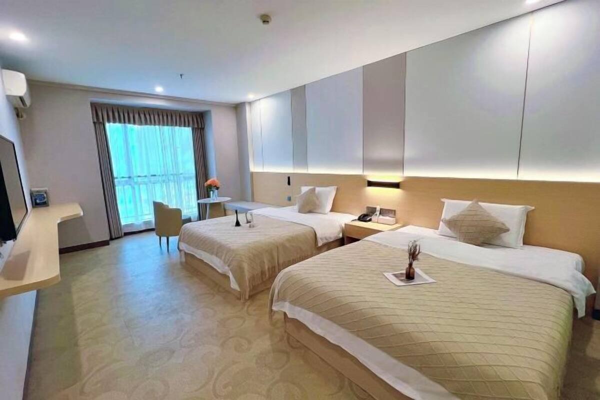 A hotel room with two beds, a desk, a chair, a window with curtains, and a wall-mounted TV.