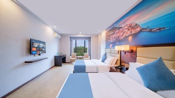 A hotel room with a bed, a desk, a chair, a TV, and a painting of a coastal scene. - Zhaoqing Jinyuan Hotel (Zhaoqing)