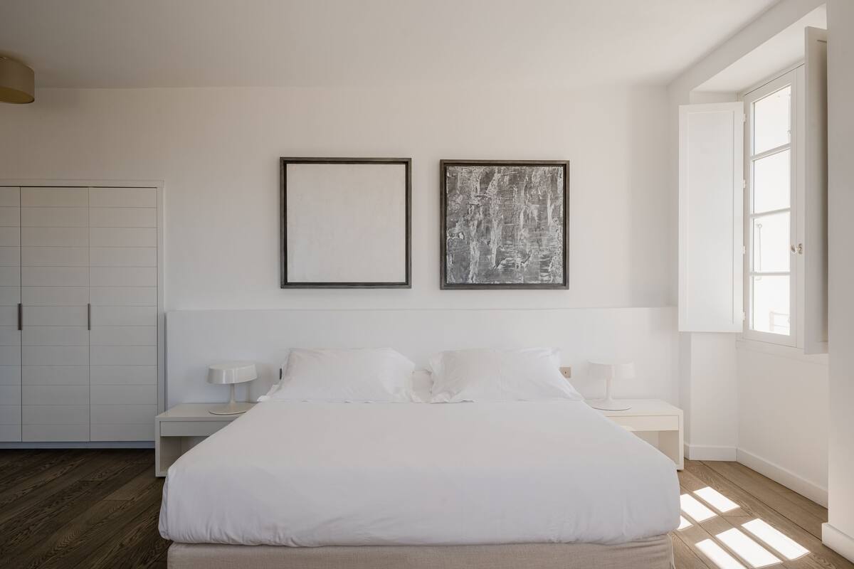 A minimalist bedroom with a large bed, two bedside tables with lamps, a wardrobe, and two framed pictures on the wall.