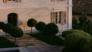 Un edificio de varios pisos con arquitectura simétrica, con ventanas y contraventanas uniformemente distribuidas, una entrada central y un área ajardinada circundante con arbustos podados.