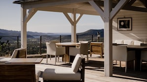 Un patio al aire libre con mesas y sillas de madera, una pérgola blanca y vistas a montañas al fondo.