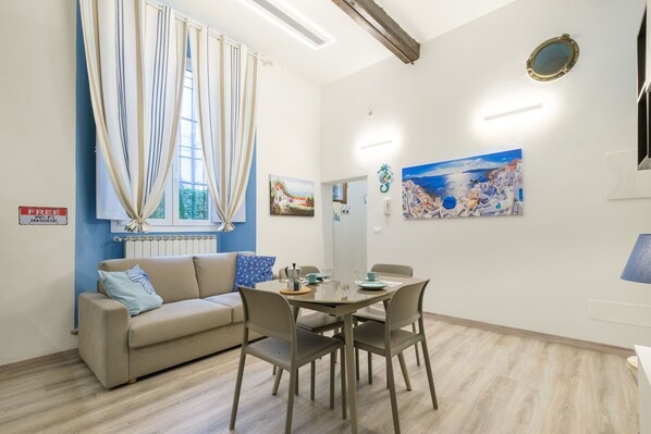 A room with a sofa, dining table, chairs, a window with curtains, and wall art.