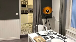 A room with a dining table set for two, a floor lamp, and a view into a kitchen area. - Le Bonnie- Ground floor studio for 2 persons (Honfleur)