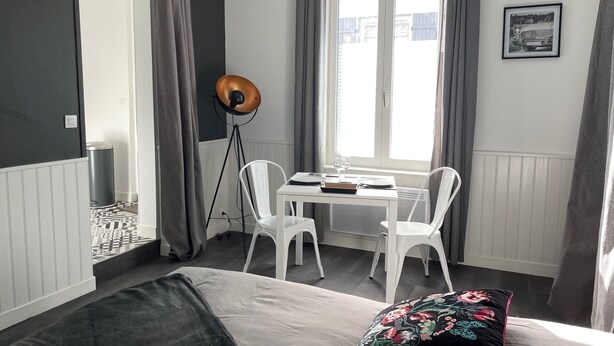 A bedroom with a bed, a desk, two chairs, a floor lamp, and a window with curtains. - Le Bonnie- Ground floor studio for 2 persons (Honfleur)