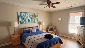 Een slaapkamer met een bed, nachtkastjes, een plafondventilator, een schilderij, een stoel, een dressoir en een raam.
