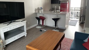 Een moderne keuken met witte kasten, een rode koelkast en een magnetron. Er staat een houten eettafel met twee barstoelen. Een televisie is aan de muur bevestigd.