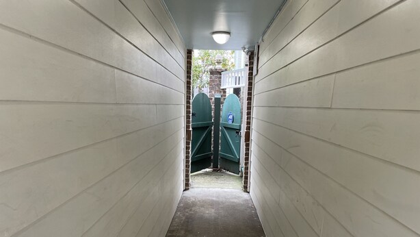 Een smalle gang met witte wanden en een betonnen vloer, die leidt naar een groene poort met een blauw bord.