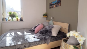 A bedroom with a bed, a window with books and flowers, a painting on the wall, and a small shelf with plants. - Captains-TERRACE- Photovoltaic- Fireplace-large quiet terrace (Göhren-Lebbin)