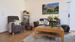 A living room with two grey armchairs, a wooden coffee table, a small table with a vase of flowers, a painting of a castle, and a floor lamp. - Captains-TERRACE- Photovoltaic- Fireplace-large quiet terrace (Göhren-Lebbin)