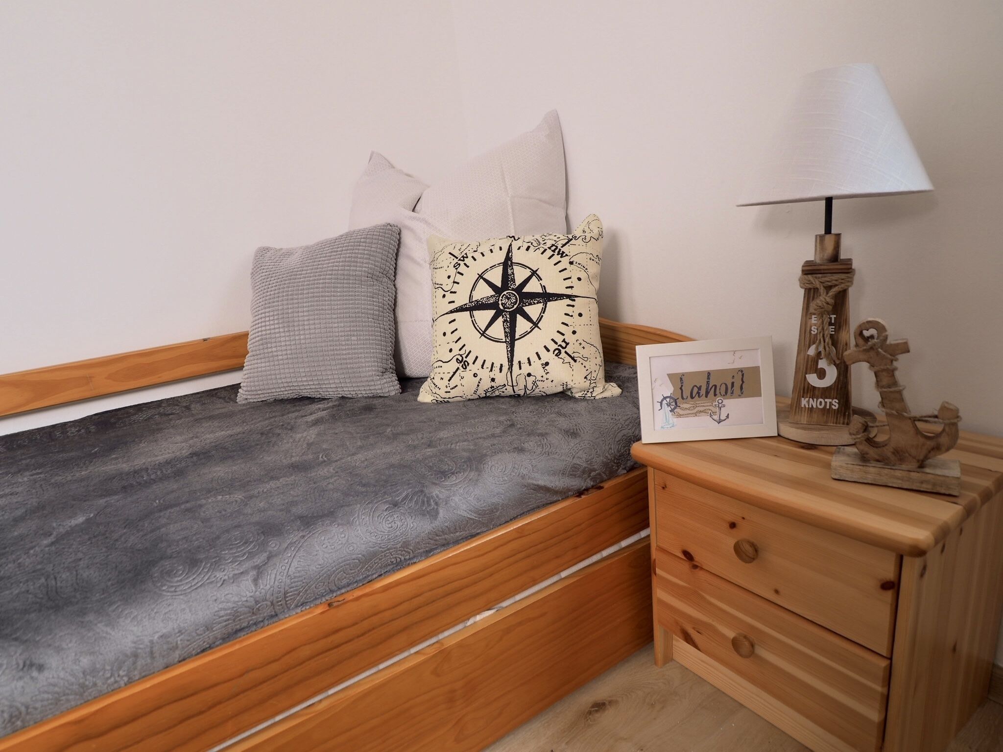 Un petit lit en bois avec une couverture grise, deux oreillers, un coussin décoré d’un motif de boussole, un tableau encadré, une table de chevet en bois avec des tiroirs et une lampe.