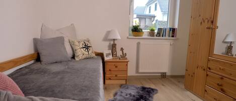 Un dormitorio con cama, cómoda de madera, ventana con vista a casas, radiador y una alfombra pequeña.