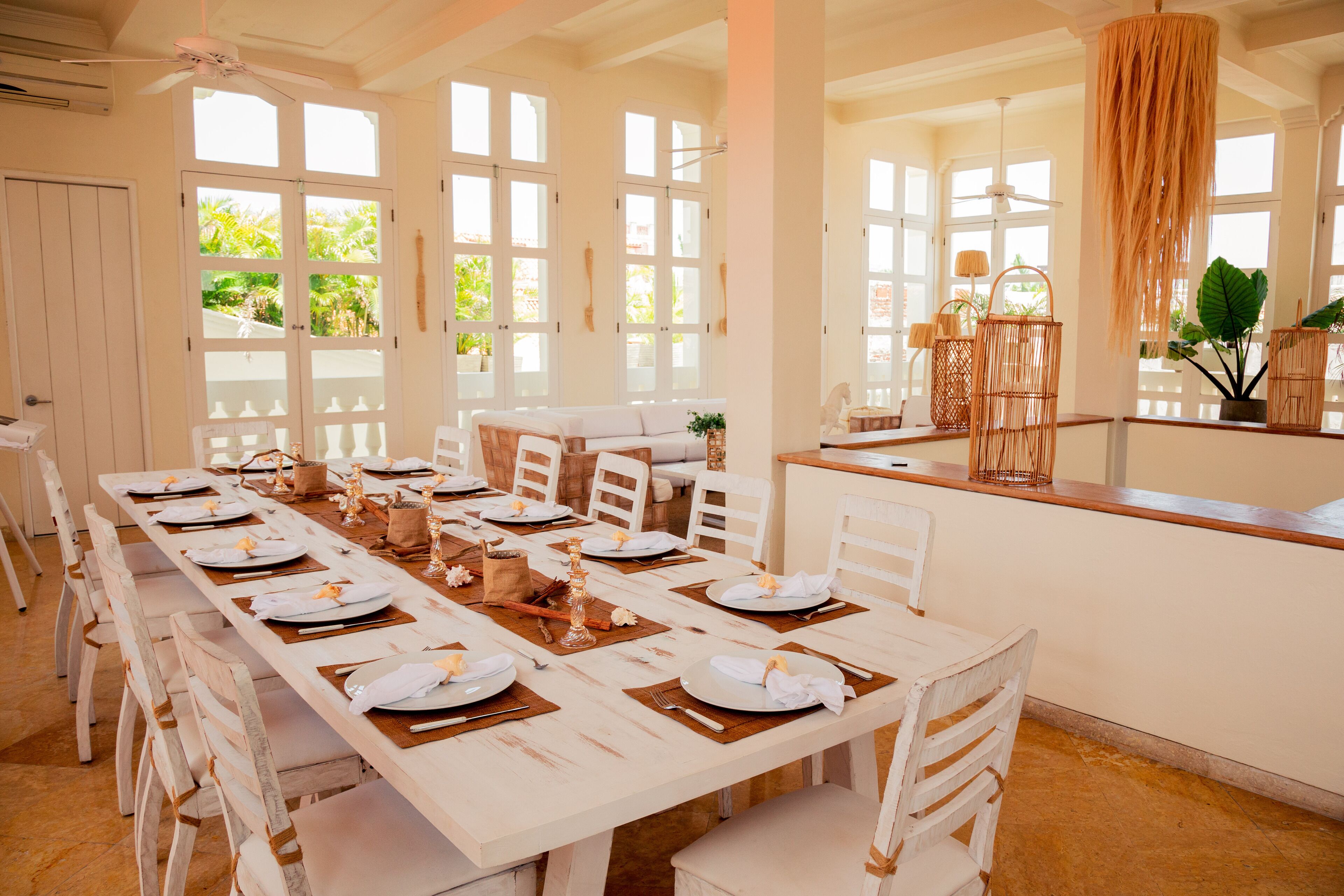 Un comedor con una larga mesa dispuesta para una comida, sillas blancas y vistas al exterior a través de amplios ventanales.