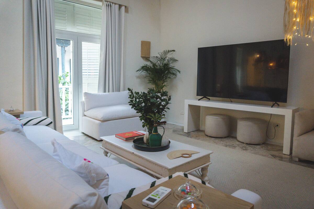 Ein Hotelzimmer mit einer Couch, einem Couchtisch mit Fernbedienung, einem Fernseher und einer Pflanze.