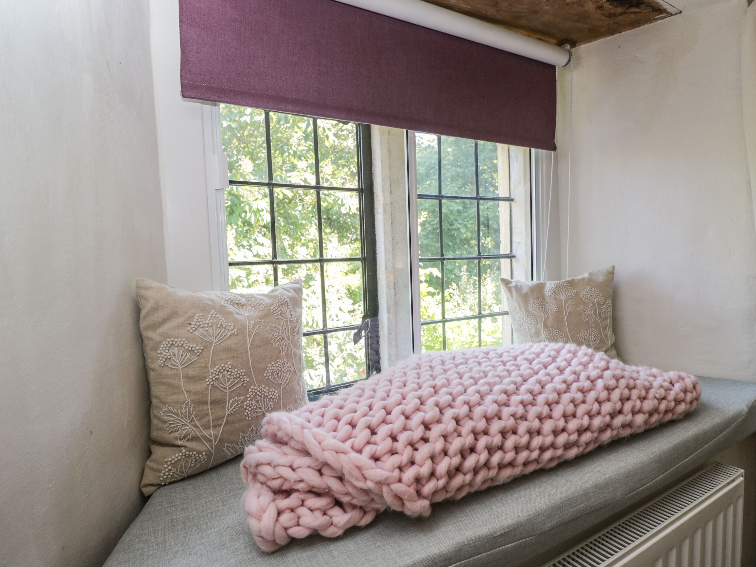 Ein Fensterplatz mit einer rosa Strickdecke, zwei Kissen mit Blumenstickerei und einem violetten Vorhang.