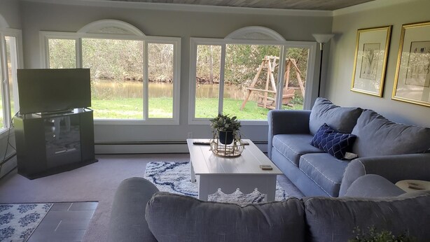 Un salón con un sofá gris, una mesa de centro y un televisor sobre un mueble bajo. Hay ventanas amplias con vistas a un jardín y un columpio.