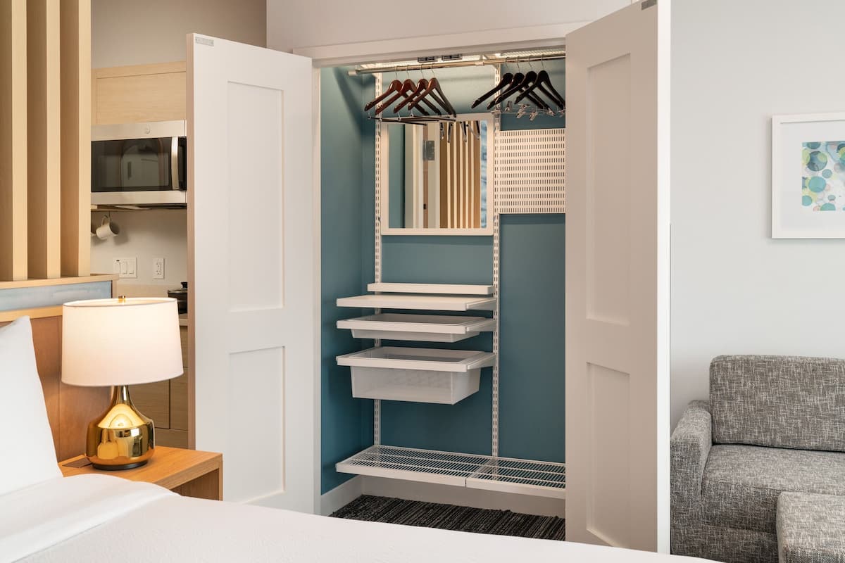 A hotel room with a walk-in closet, a bed, a bedside lamp, a microwave, and a grey armchair.