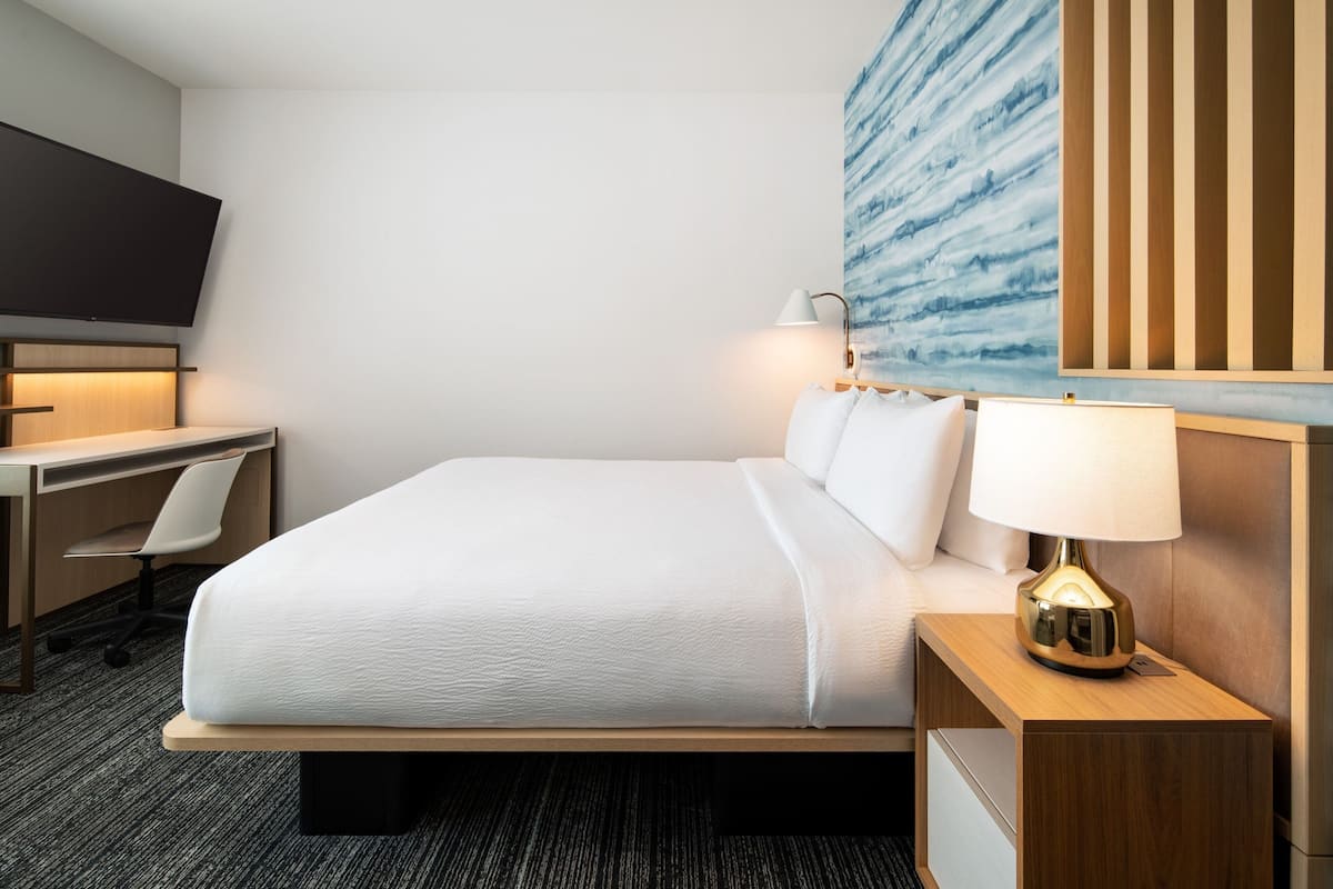 A hotel room with a bed, bedside table, lamp, desk, chair, and a TV mounted on the wall.