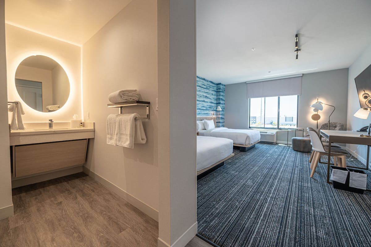A modern hotel room with a bathroom featuring a round mirror, a sink, and a towel rack. The room includes a bed, a desk with a chair, and a large window offering a view.