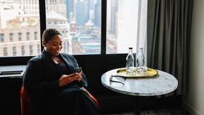 A person seated by a window with a city view, a small table with bottles and glasses, and a pair of glasses on the table. - voco Chicago Downtown - Riverwalk by IHG (Chicago)