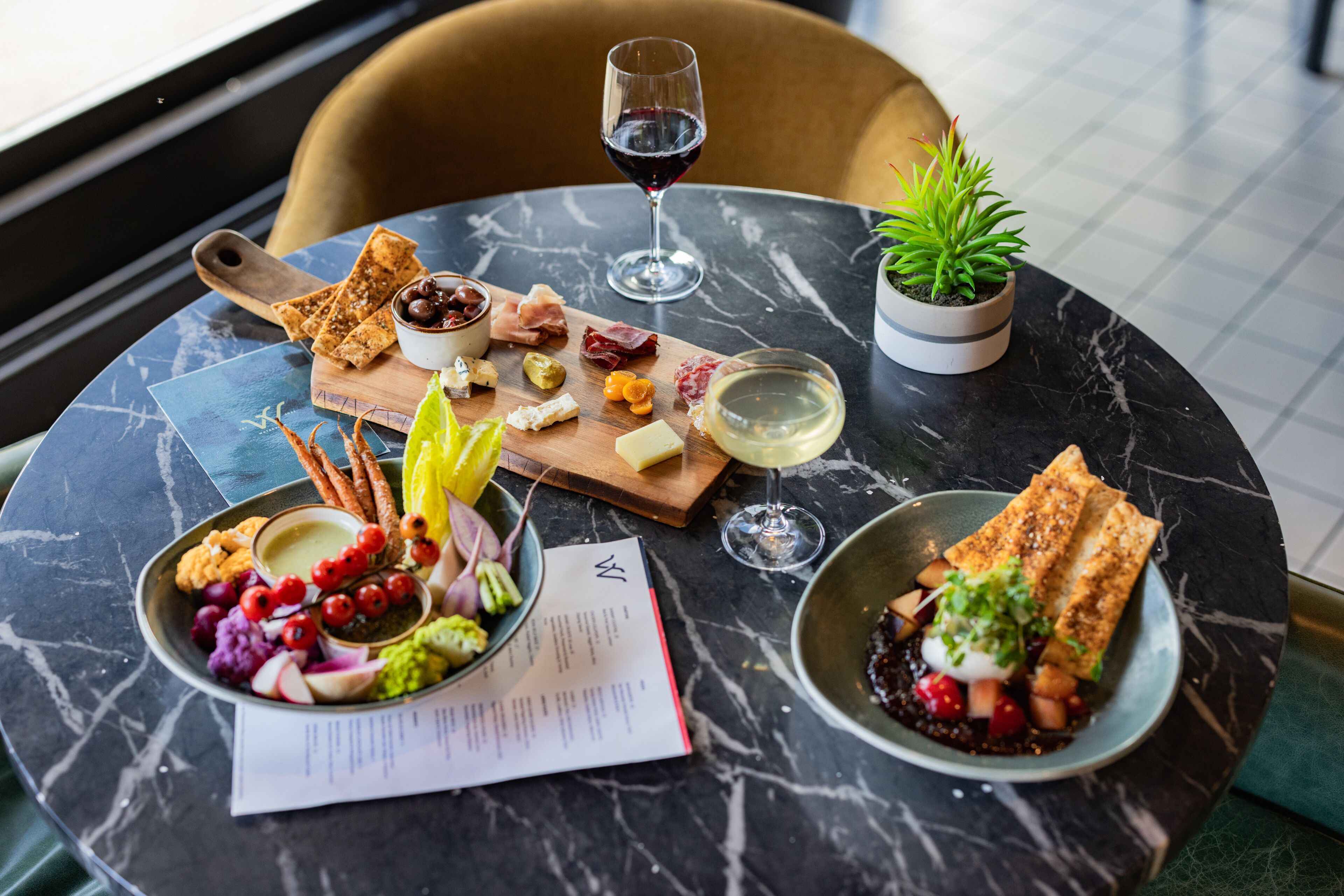 A marble table with a variety of food items, including cheese, fruits, and bread, accompanied by wine glasses and a menu.