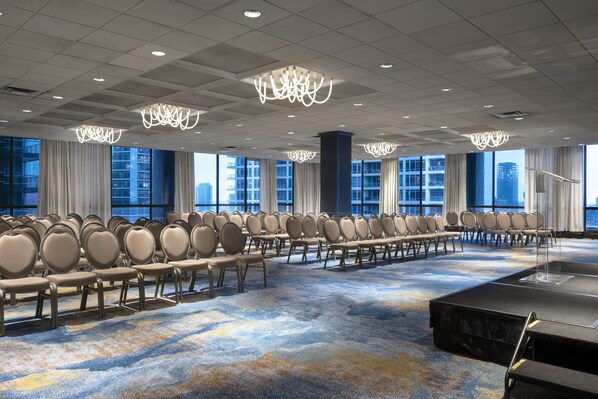A conference room with rows of chairs, a stage, and large windows with a city view.