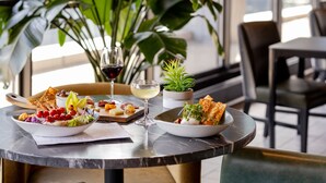 Una mesa de comedor con ensalada, queso y vino, rodeada de sillas y una planta grande.