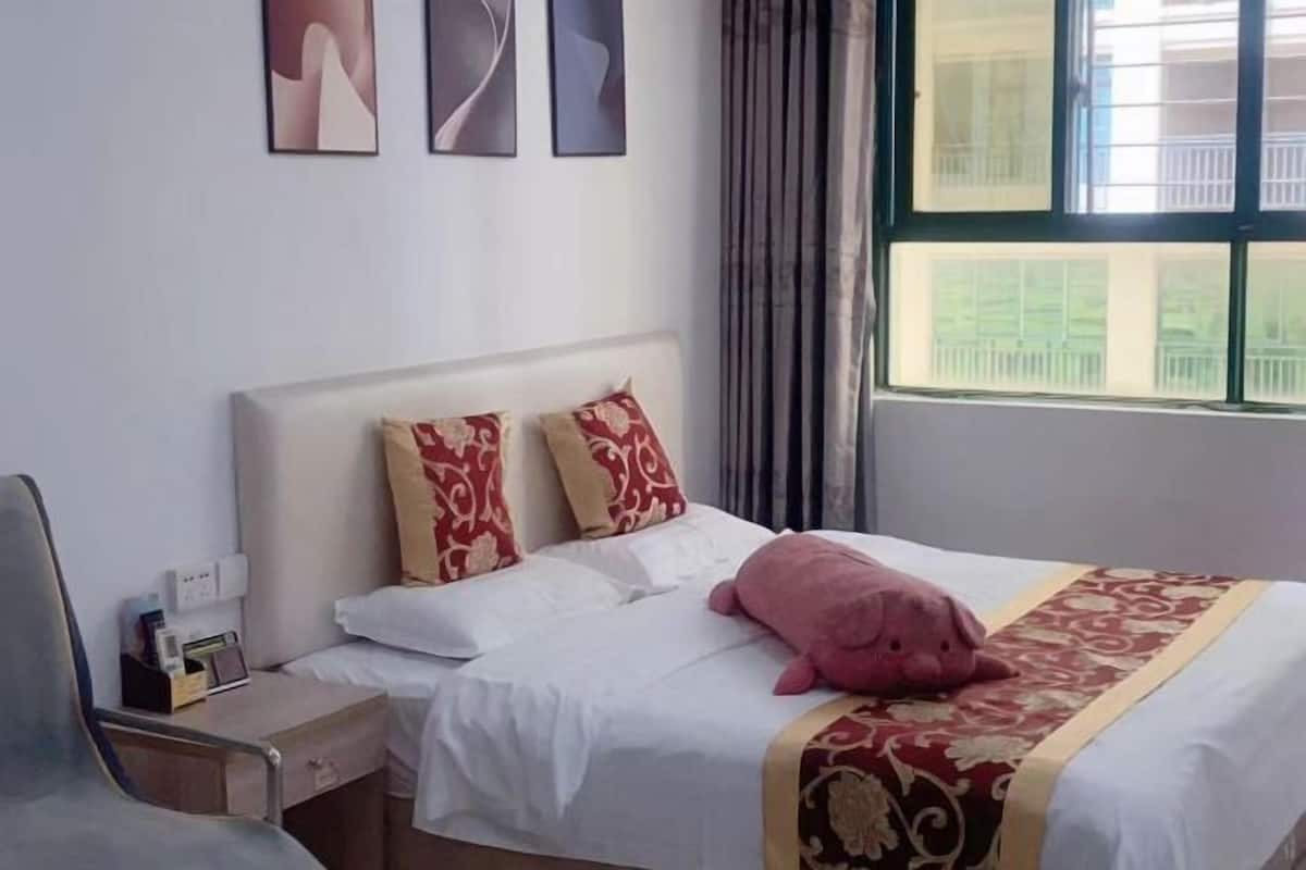 A hotel room with a bed, pillows, a bedside table with a lamp, a window with curtains, and artwork on the wall.