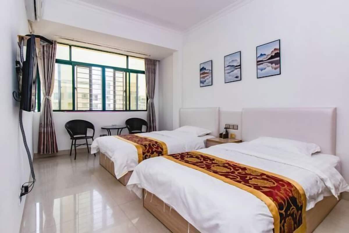 A hotel room with two beds, a window with curtains, a chair, a small table, and artwork on the wall.