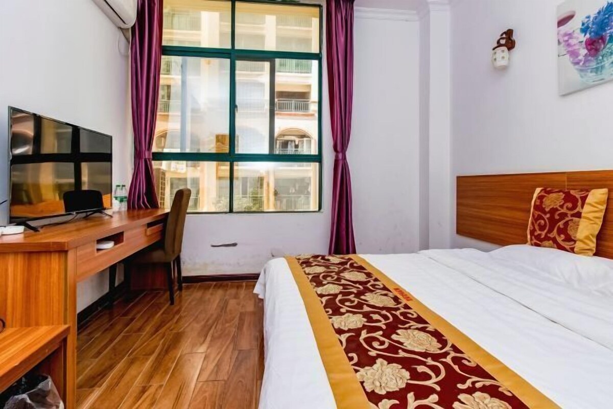 Ein Hotelzimmer mit einem Holztisch, einem Bett mit gemustertem Überwurf, einem Fernseher und einem Fenster mit Vorhängen.