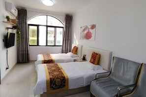 Two single beds with white linens and decorative pillows, a grey armchair, and a large window with an outside view.