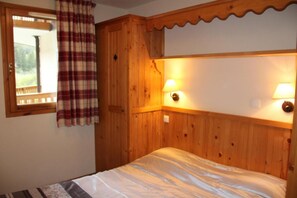 A wooden room with a bed, two wall-mounted lamps, a window with a plaid curtain, and a view of greenery outside. - 150m from shops, 2nd floor, view swimming pool, fitness, swimming pool, balcony, tv, ski locker (Lanslebourg-Mont-Cenis)