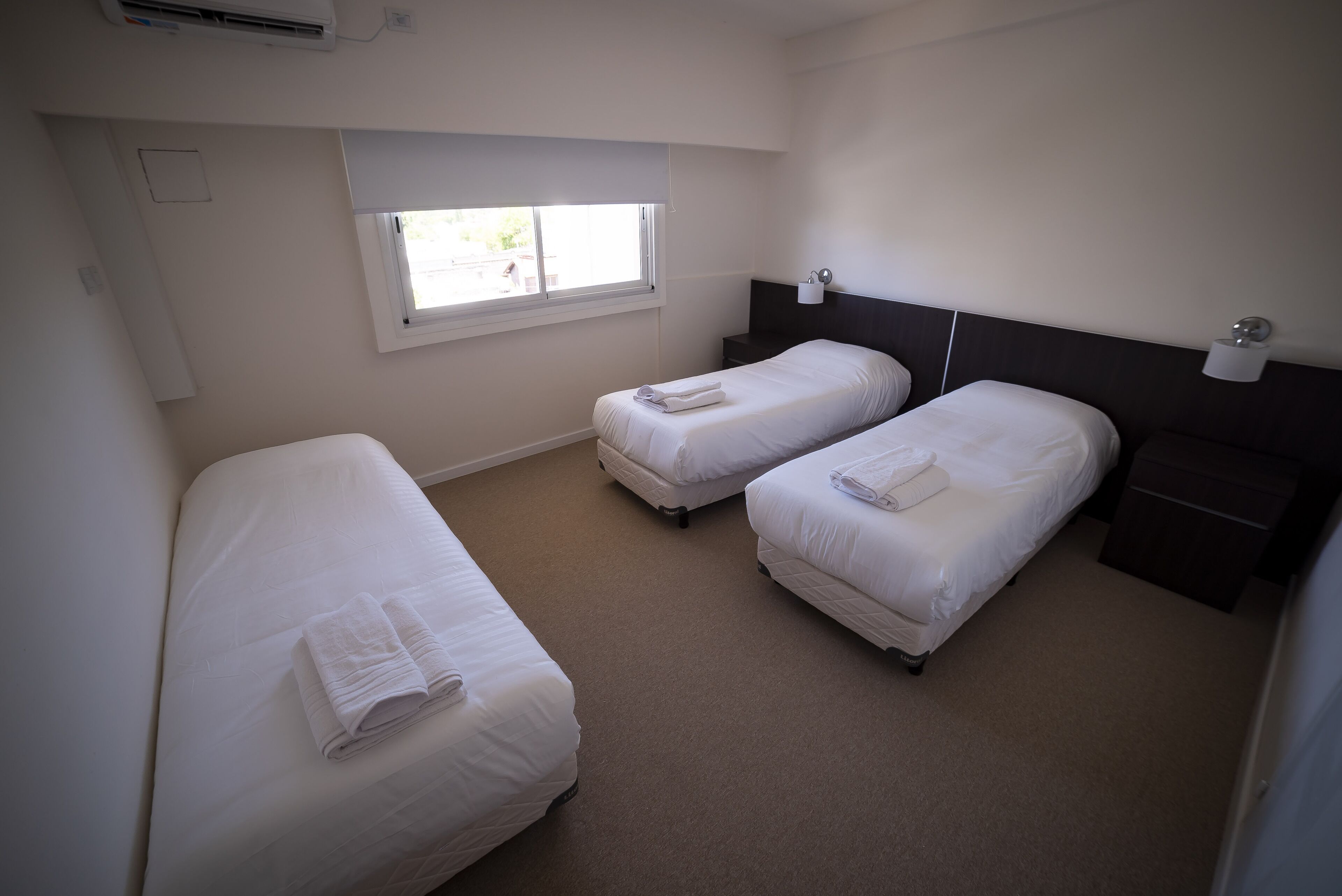 A hotel room with two single beds, a window, a bedside table, and a wall-mounted lamp.